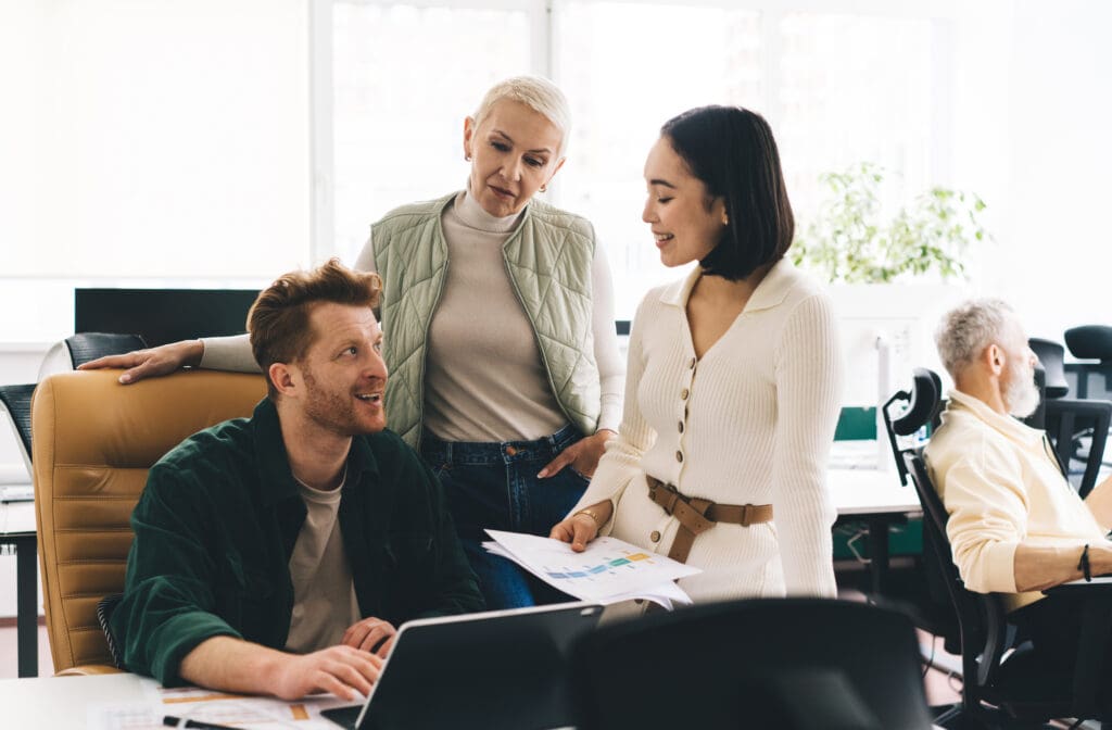 Group of multiracial colleagues discussing office work near table of sitting male with laptop while Asian female employee standing with documents and looking at each other.